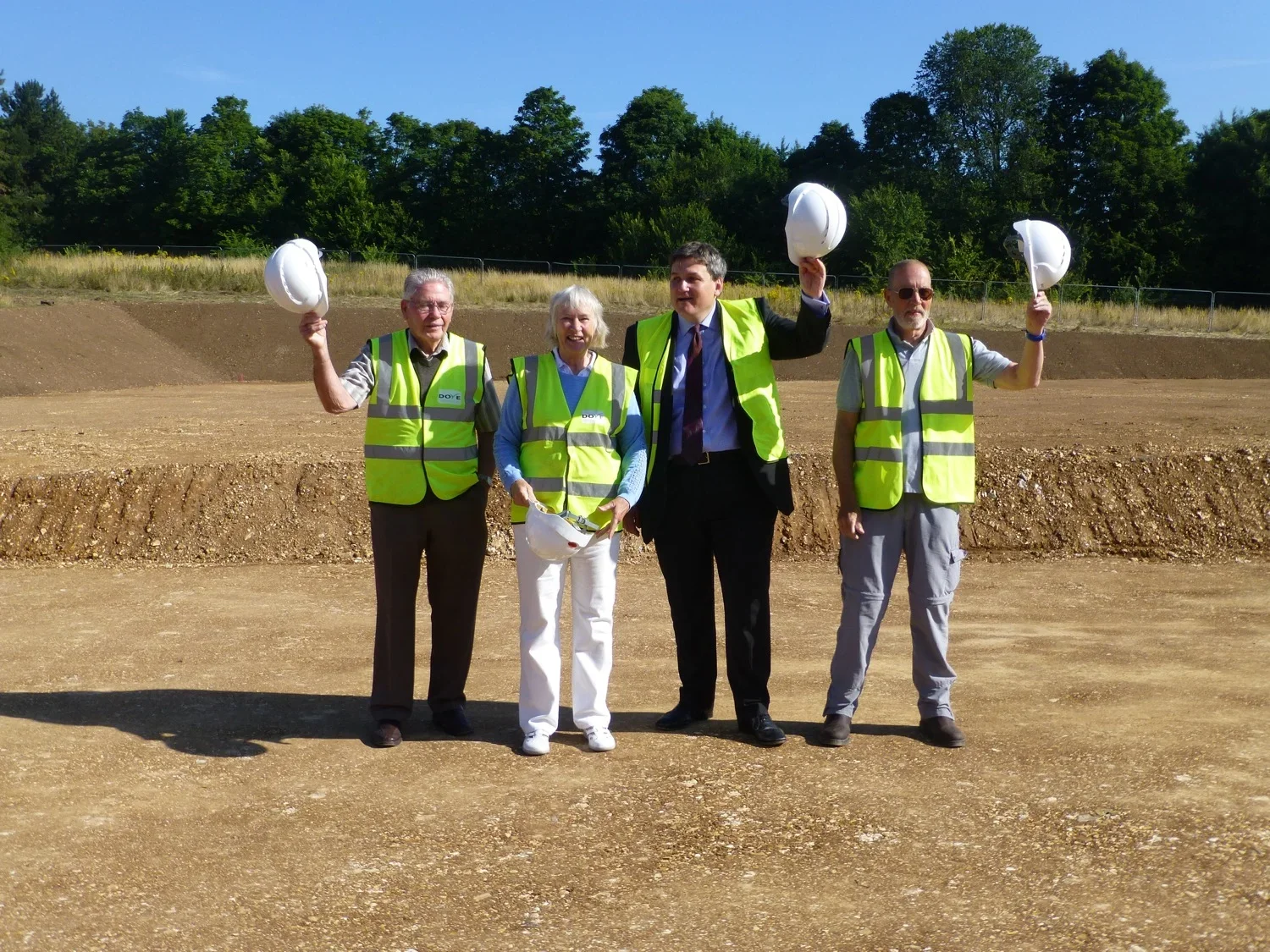 Four people in safety vests holding white construction helmets, standing on a dirt construction site with trees in the background during daytime.