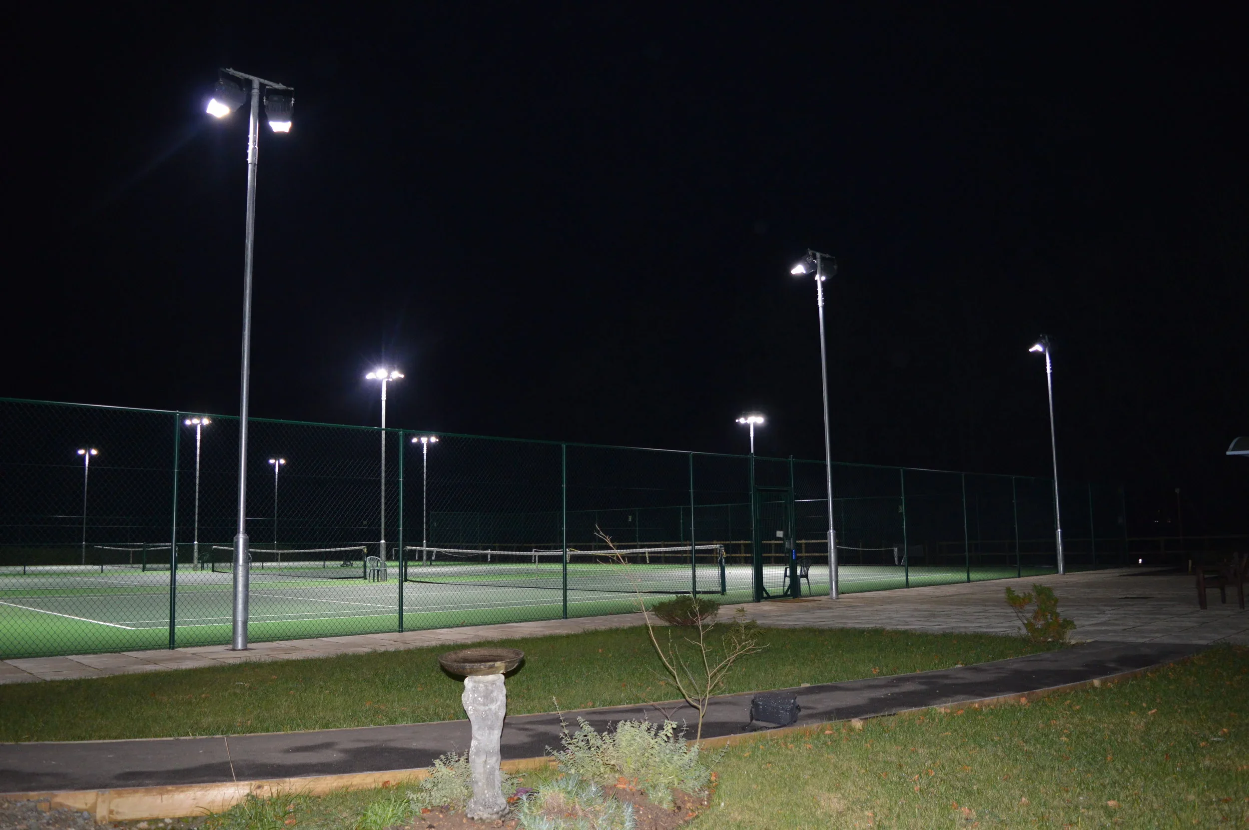Tennis courts illuminated at night with multiple tall lights, surrounded by a fence, with sidewalk and grass in the foreground.