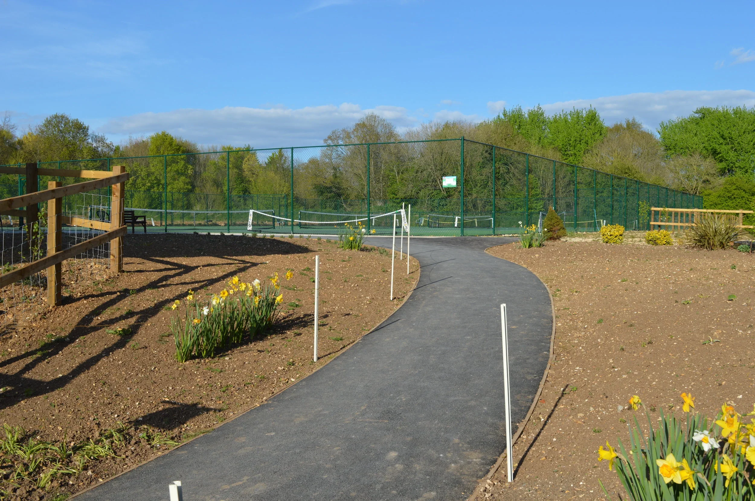 A pathway leading to a tennis court surrounded by greenery and flower beds.