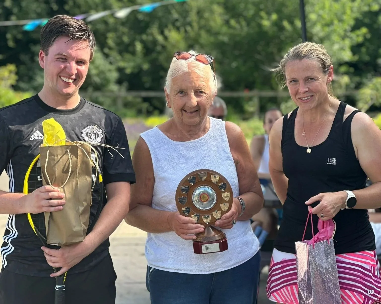 Three women smiling, one holding a trophy and gifts, standing outdoors with trees in the background.