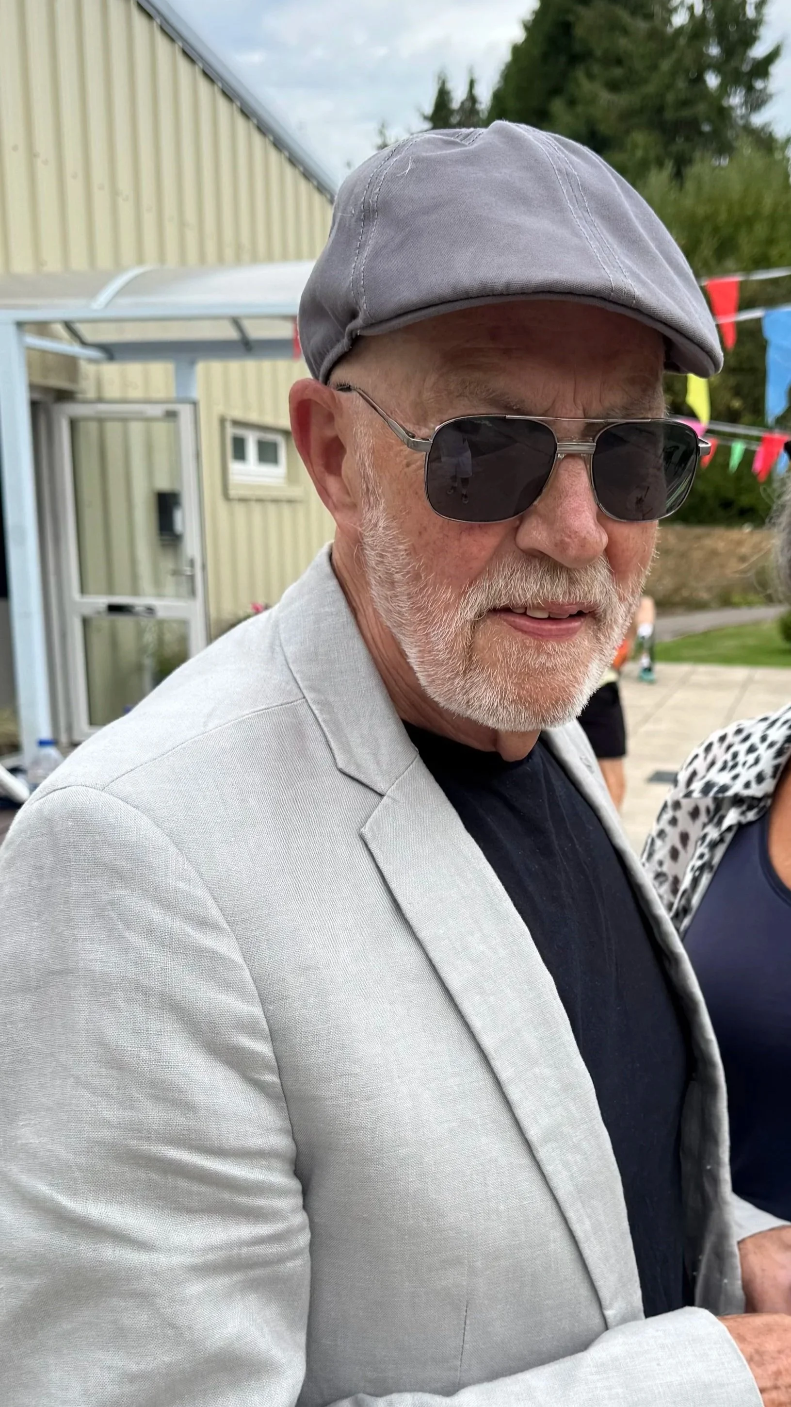 An older man with a white beard and glasses, wearing a gray hat and a light gray blazer, standing outdoors near a building with colorful banners in the background.