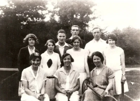 Black and white photo of nine people, six women and three men, posing outdoors with trees in the background, possibly from the early 20th century.