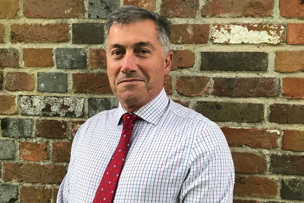 A middle-aged man with short gray and black hair, wearing a white checkered shirt and a red tie, standing against a brick wall background.