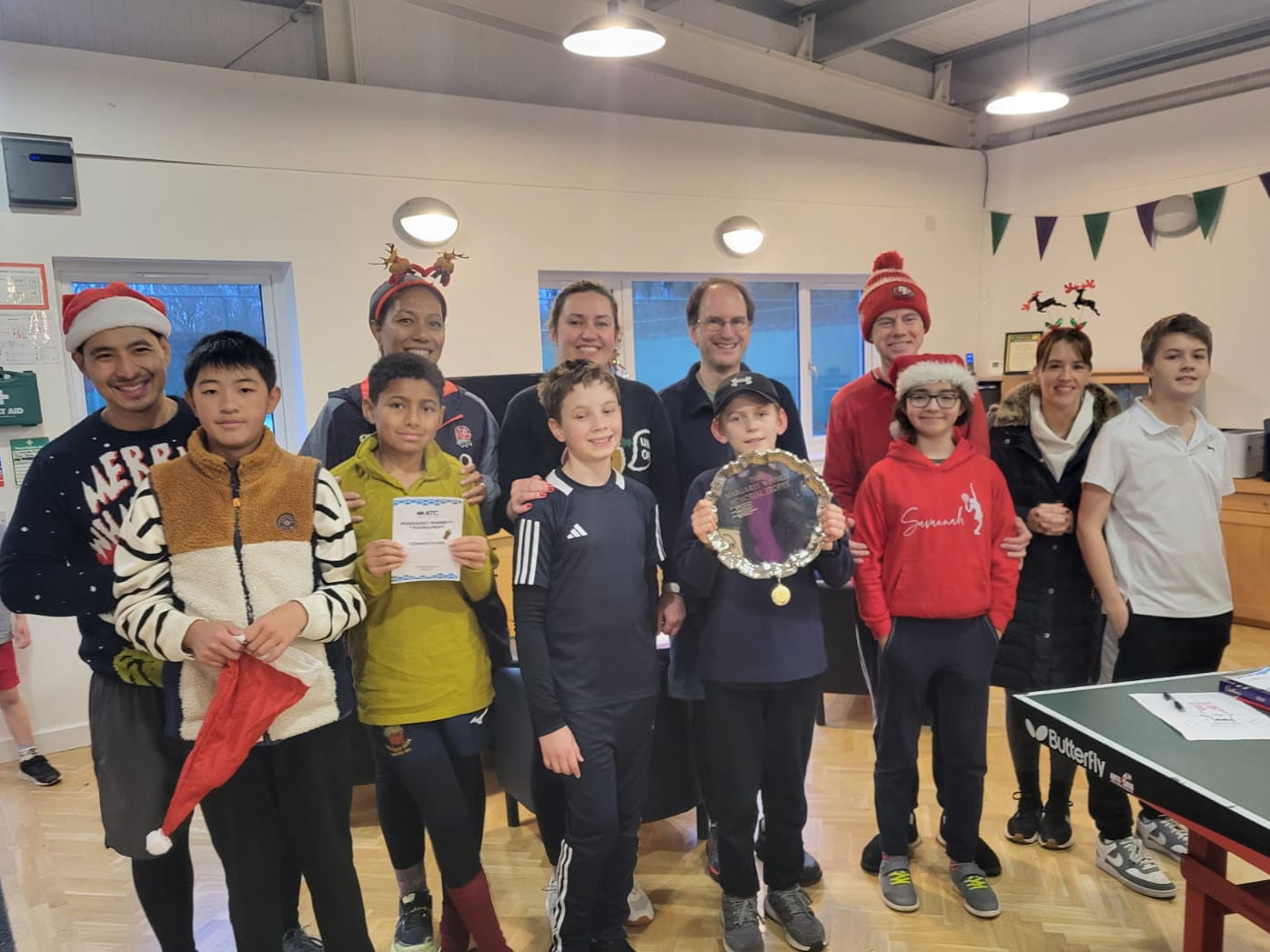 Group of children and adults celebrating, some wearing Christmas-themed clothing and accessories, in an indoor setting with holiday decorations and a ping pong table.