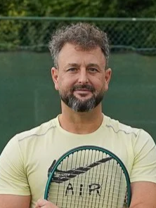 Man with curly hair and beard holding a tennis racket on a tennis court