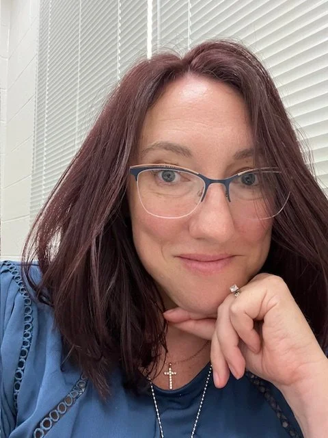 Profile picture of white lady with medium length auburn hair, blue eyes, and glasses. She is resting her hand against her chin and has a closed-mouthed smile. Vertical white blinds in the background
