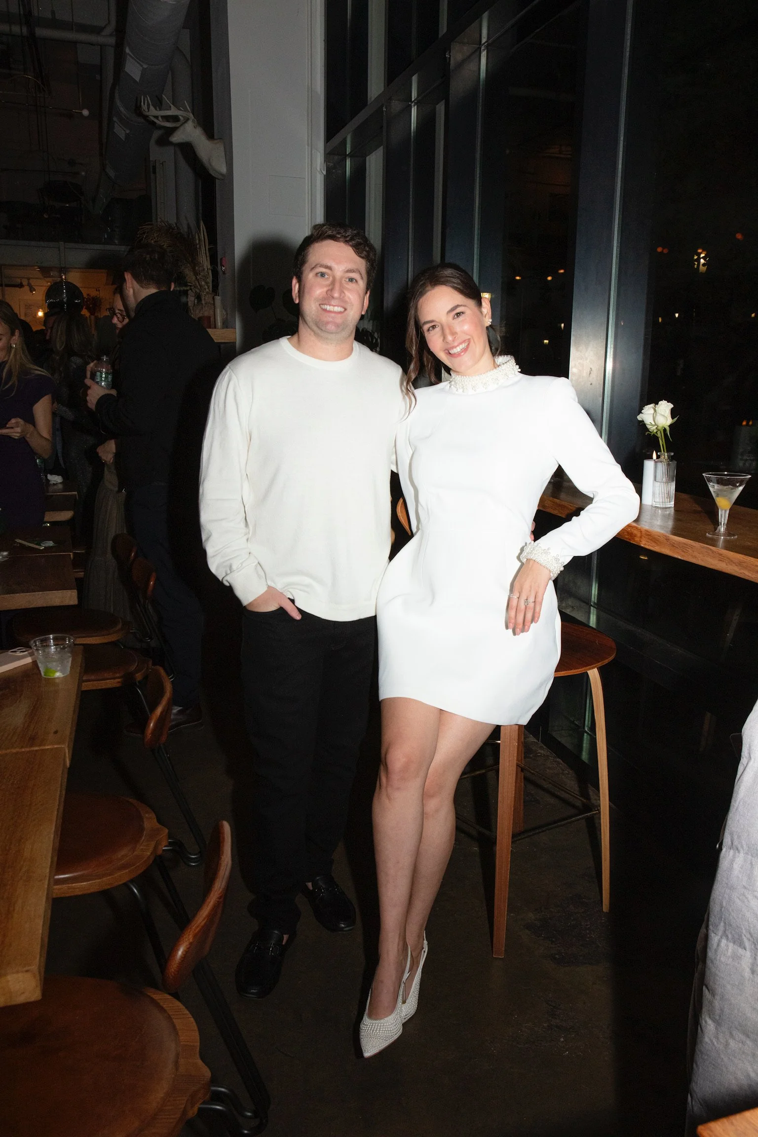 A man and woman smiling at a social gathering in a restaurant or bar. The woman is wearing a white dress and heels, while the man is dressed casually in a white sweater and black pants.