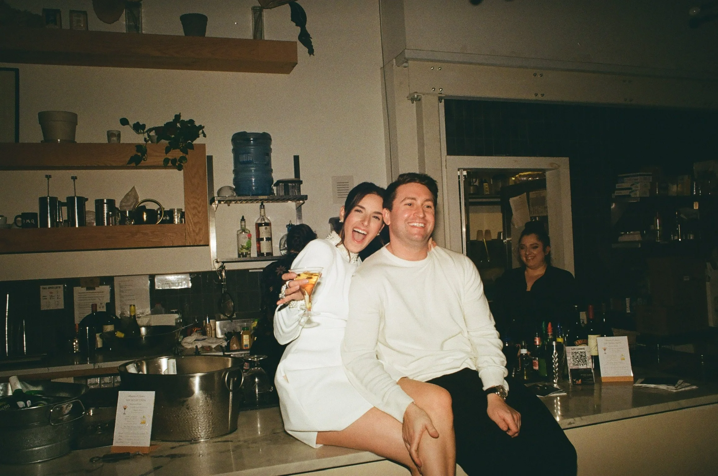 A woman and a man sitting together on a restaurant counter, smiling and enjoying drinks. The woman is holding a glass of champagne, wearing a white dress, with her arm around the man, who is dressed in a white shirt. A waitress stands behind them in the background, and shelves with kitchen items are visible.