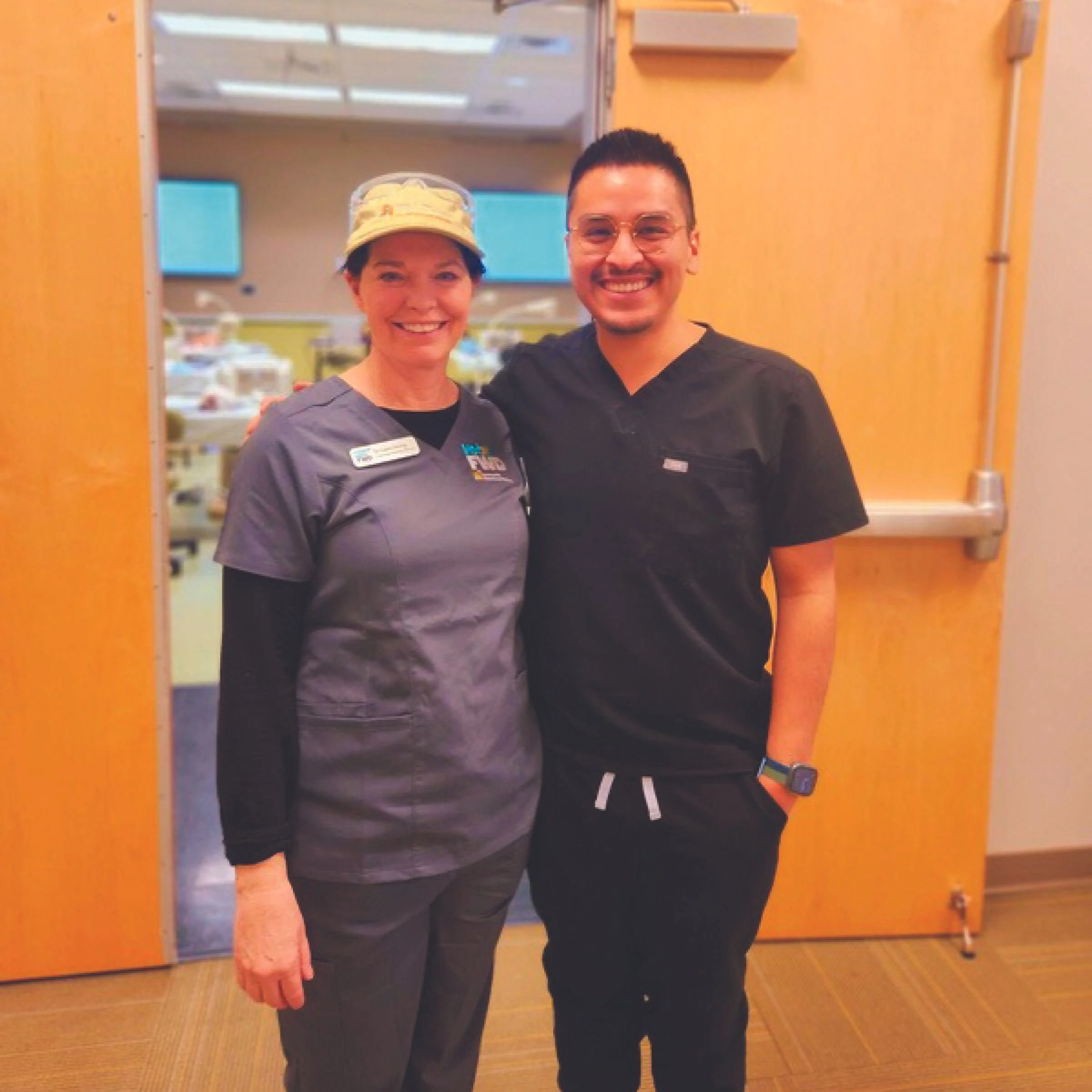 A woman and a man wearing scrubs standing side by side, smiling, in a hospital or healthcare setting.