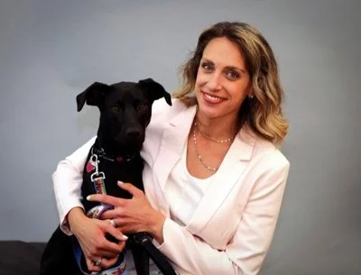 A woman with blonde hair wearing a light pink blazer smiling and holding a black dog with a pink collar against a gray background.