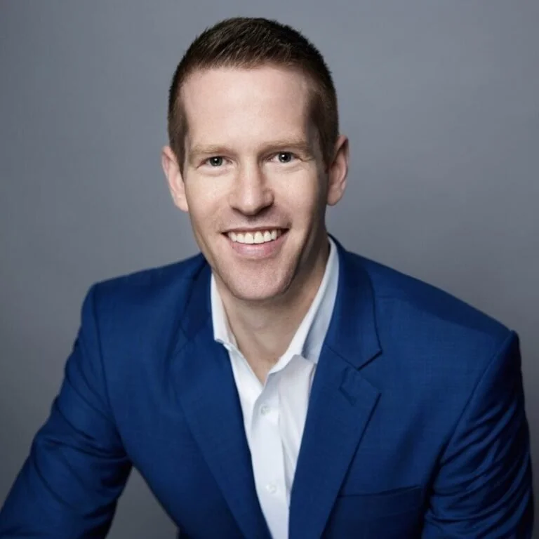 Headshot of a smiling man with short brown hair wearing a blue suit jacket and white collared shirt against a gray background.