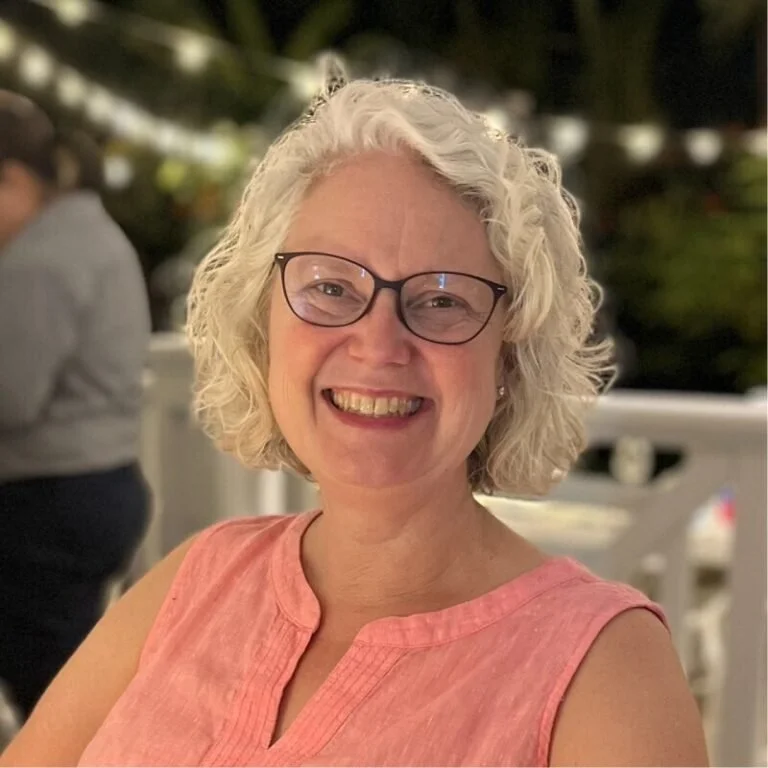 A woman with curly gray hair wearing glasses and a pink sleeveless top, smiling at the camera indoors or outdoors at night.
