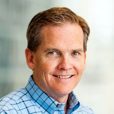 A smiling man with light brown hair, blue eyes, wearing a light blue checkered shirt, standing indoors with a blurred background.
