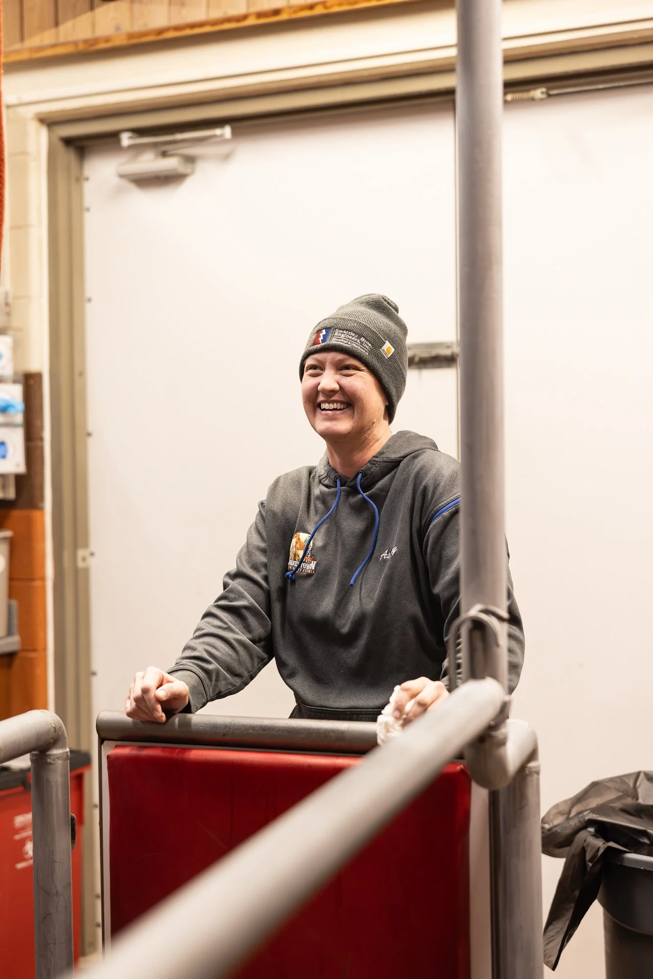 A young woman smiling, wearing a gray beanie and hoodie, standing behind a red cart in a warehouse or garage setting.