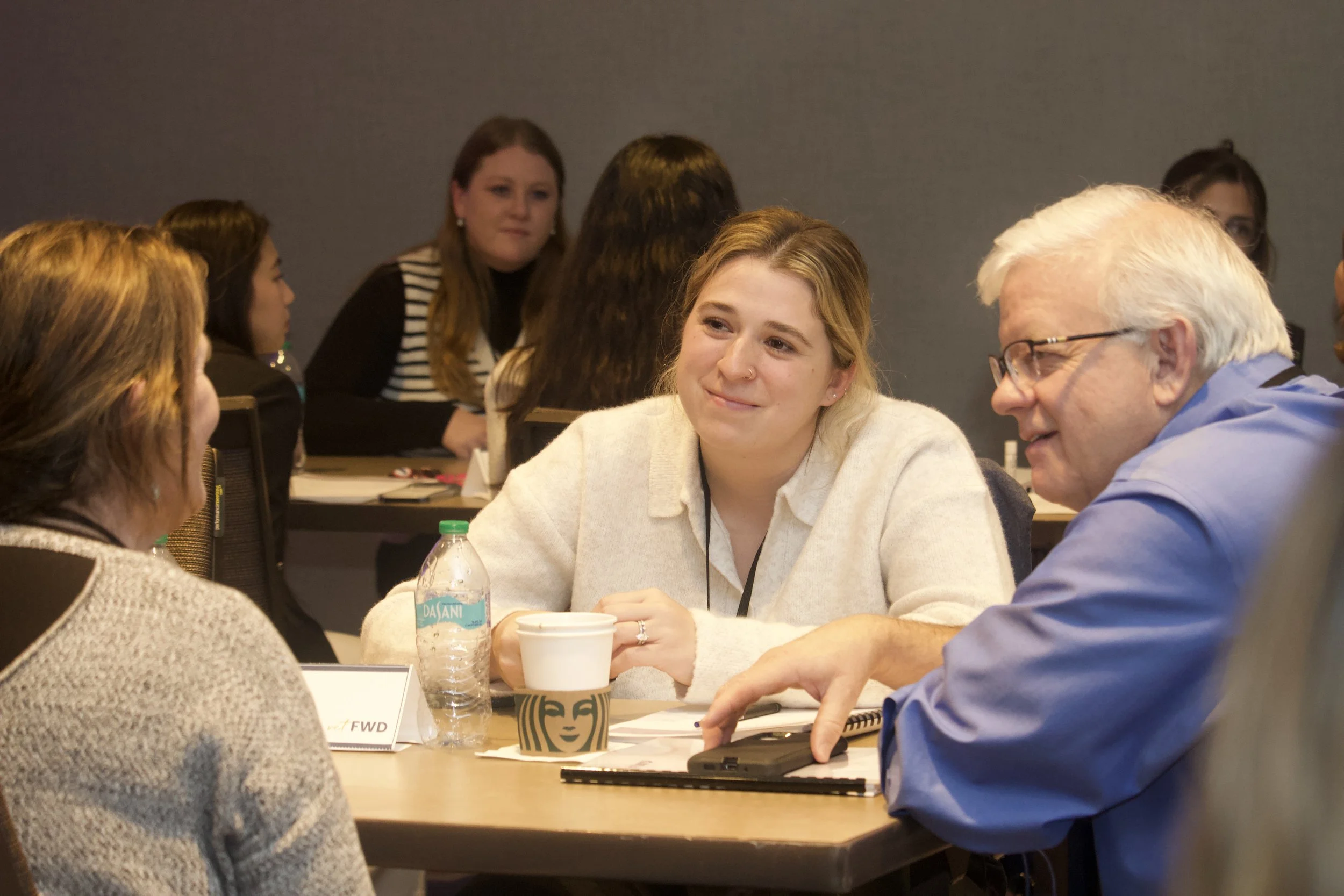 A young woman with long brown hair and a navy sweatshirt with a veterinary clinic logo, smiling slightly, with a green parrot perched on her shoulder in an indoor setting.