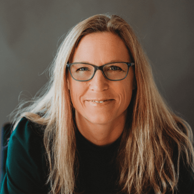 A woman with long blonde hair wearing glasses and a dark top, smiling at the camera.
