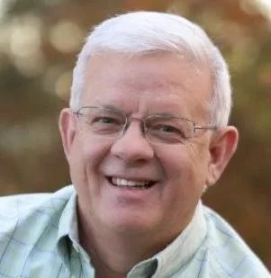 A smiling older man with white hair and glasses, wearing a light-colored shirt outdoors with blurred autumn trees in the background.