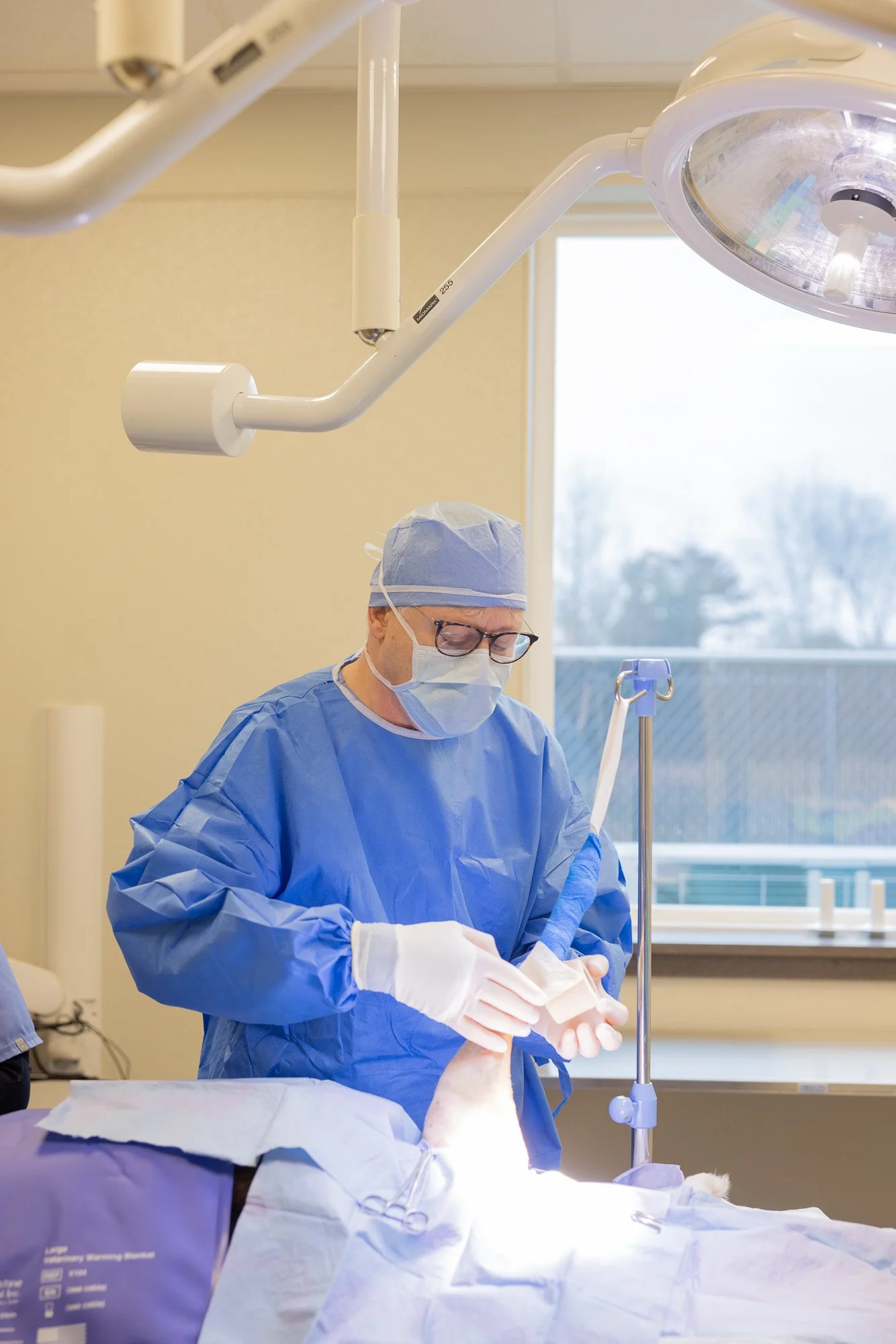 A surgeon dressed in blue scrubs, a surgical cap, mask, and gloves is performing an operation in an operating room under bright surgical lights.