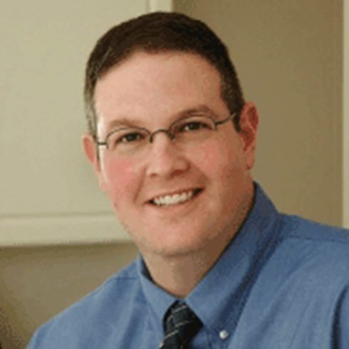 A man with short dark hair, glasses, and a friendly smile, wearing a blue shirt and a dark tie, sitting indoors near a light-colored wall.