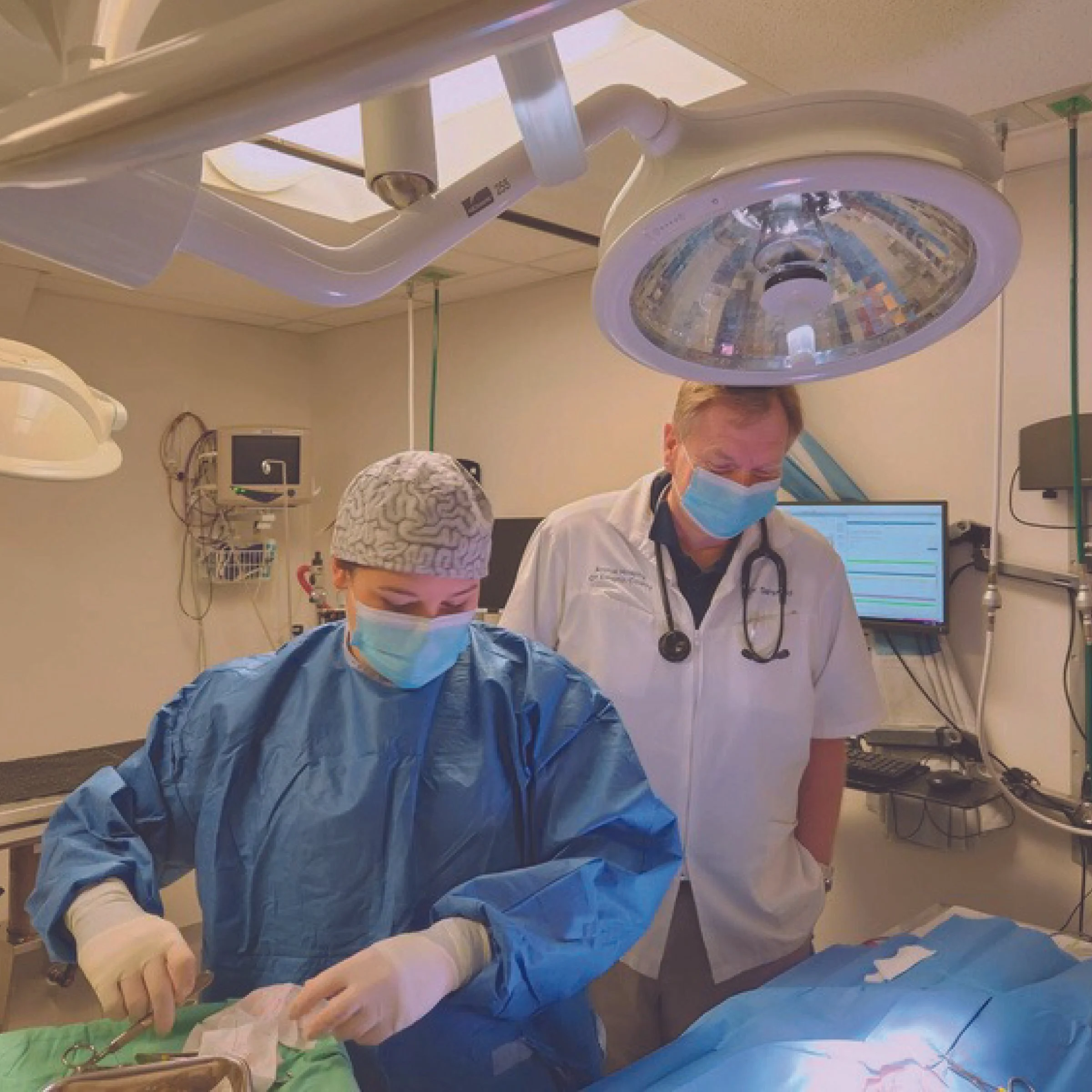 Two medical professionals, a female surgeon and a male doctor, working together in an operating room. They are wearing surgical masks, gloves, and scrubs, with surgical lights overhead. The female is focused on a procedure, while the male observes and talks. Medical equipment and monitors are visible in the background.