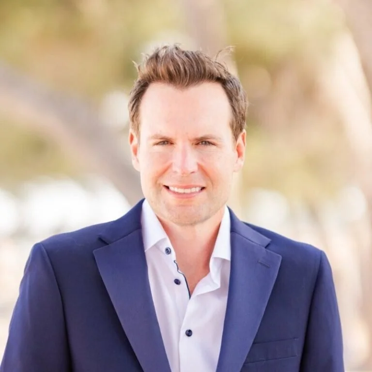 A man in a navy blue suit and white shirt smiling outdoors with blurred trees in the background.