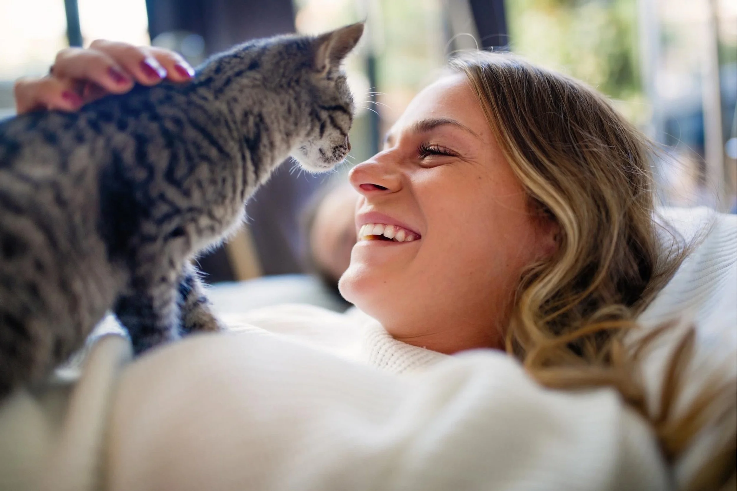 A young woman with wavy blonde hair smiling as she gently touches a gray tabby kitten's face while lying down indoors.