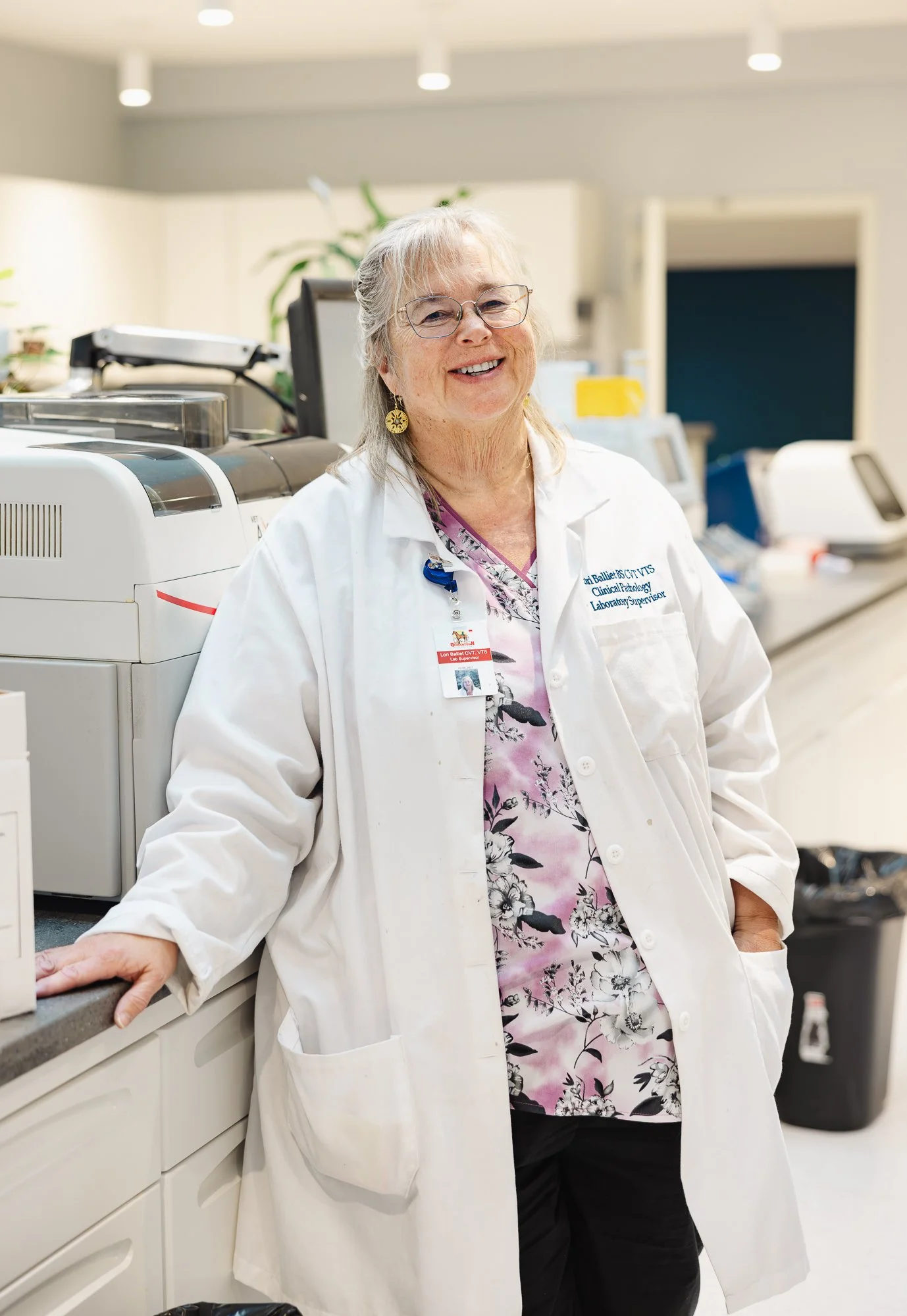 A smiling senior female healthcare professional in a white lab coat standing in a medical laboratory or clinical setting.