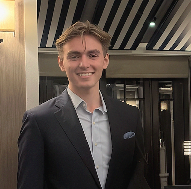 A young man in a dark suit and white shirt smiling in an indoor setting with modern decor.