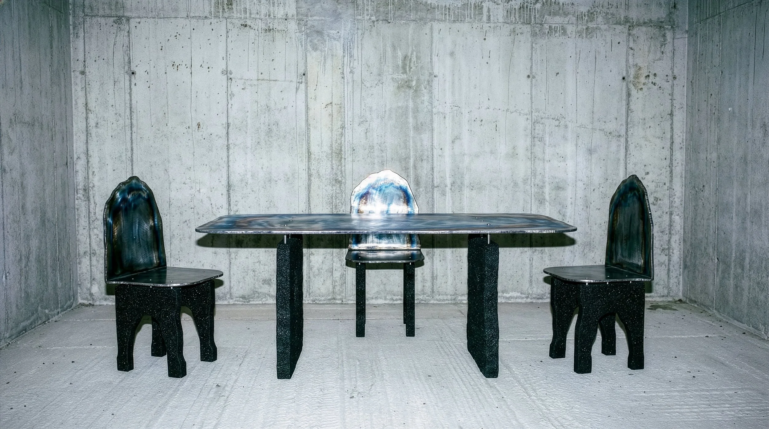 Une table et trois chaises dans une pièce en béton gris.  PAUL HARDY
