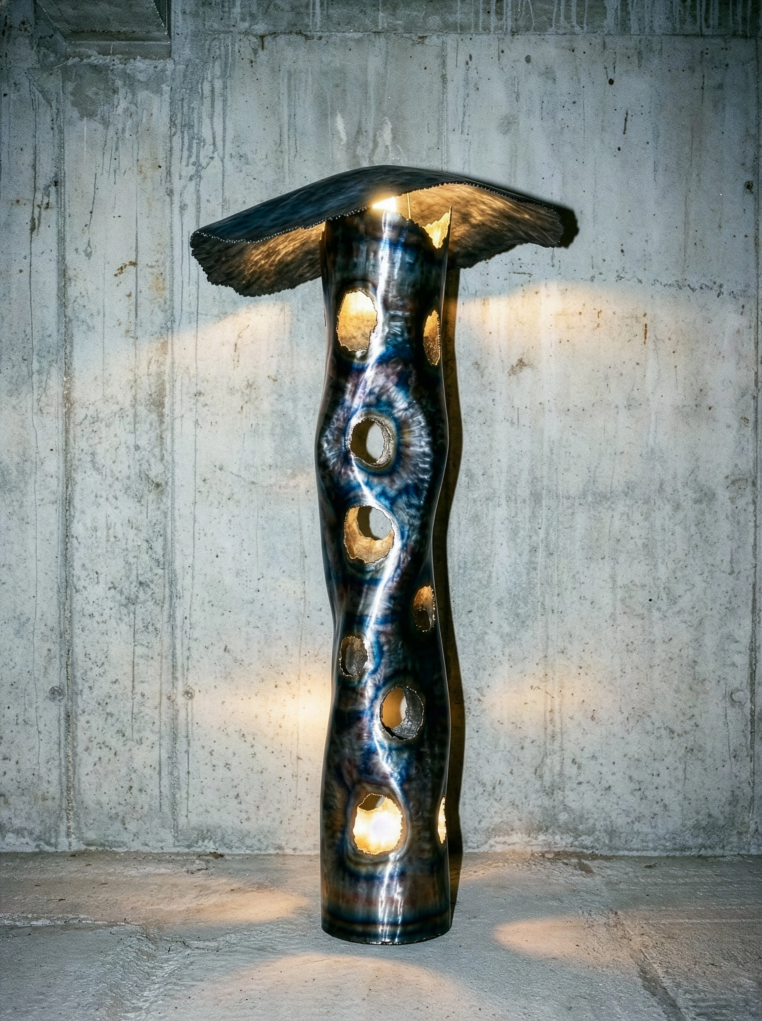 Une sculpture moderne en forme de colonne avec des trous circulaires, en matériaux métalliques ou composites, exposée contre un mur en béton.