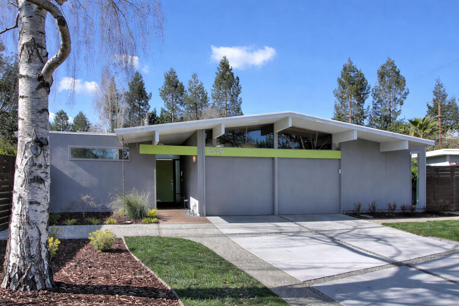 Modern house with gray exterior, green accents, and a flat roof, surrounded by a landscaped yard with trees and clear blue sky.