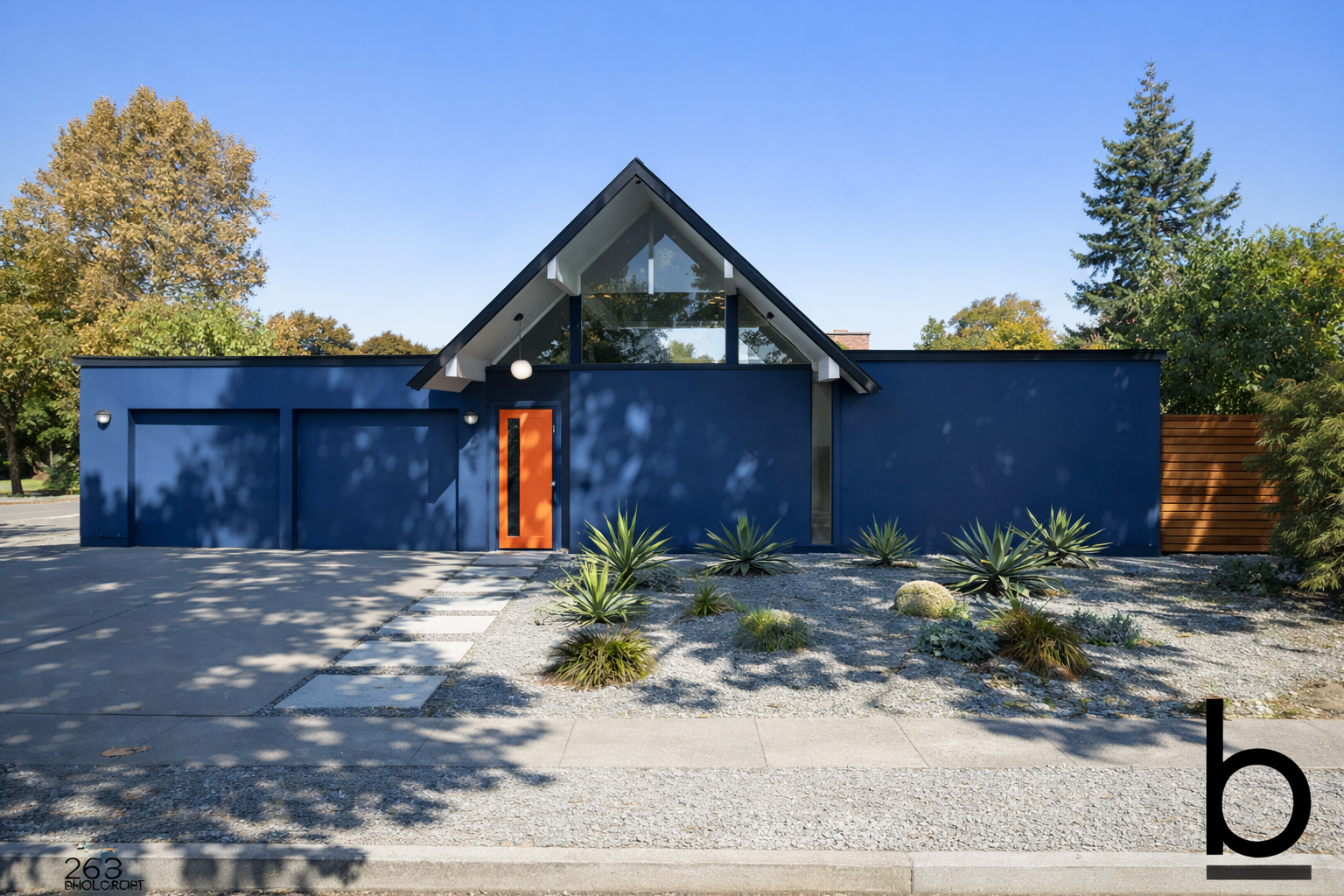 Front view of a modern blue house with a steep triangular glass roof and an orange front door, surrounded by desert plants and trees.