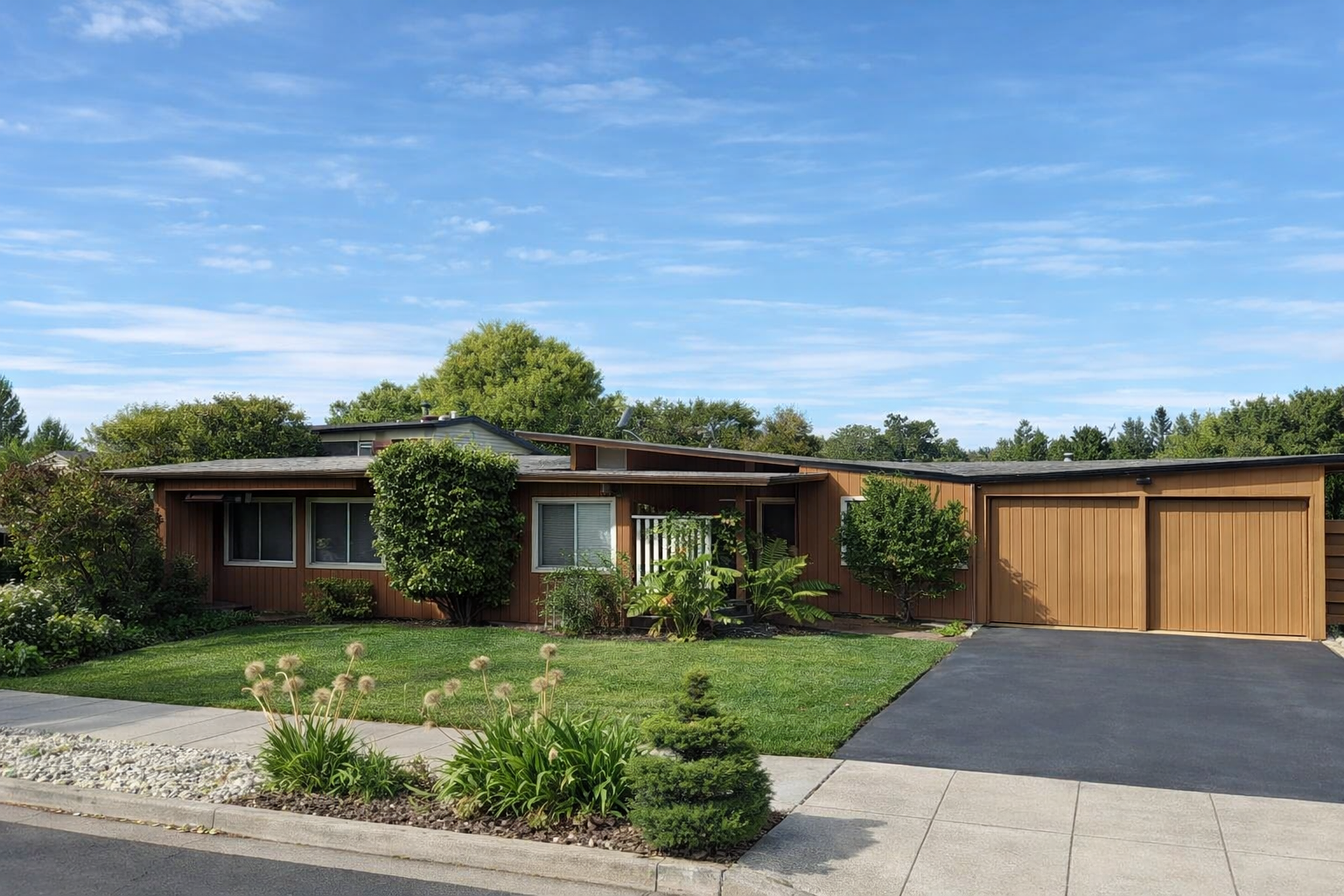 Modern single-story house with wooden exterior, well-maintained lawn, trees, and a driveway under a blue sky.