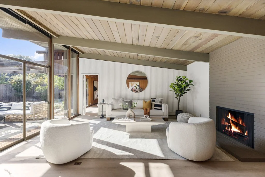 Modern living room with white and beige furniture, a fireplace, large glass sliding doors, and a green potted plant.