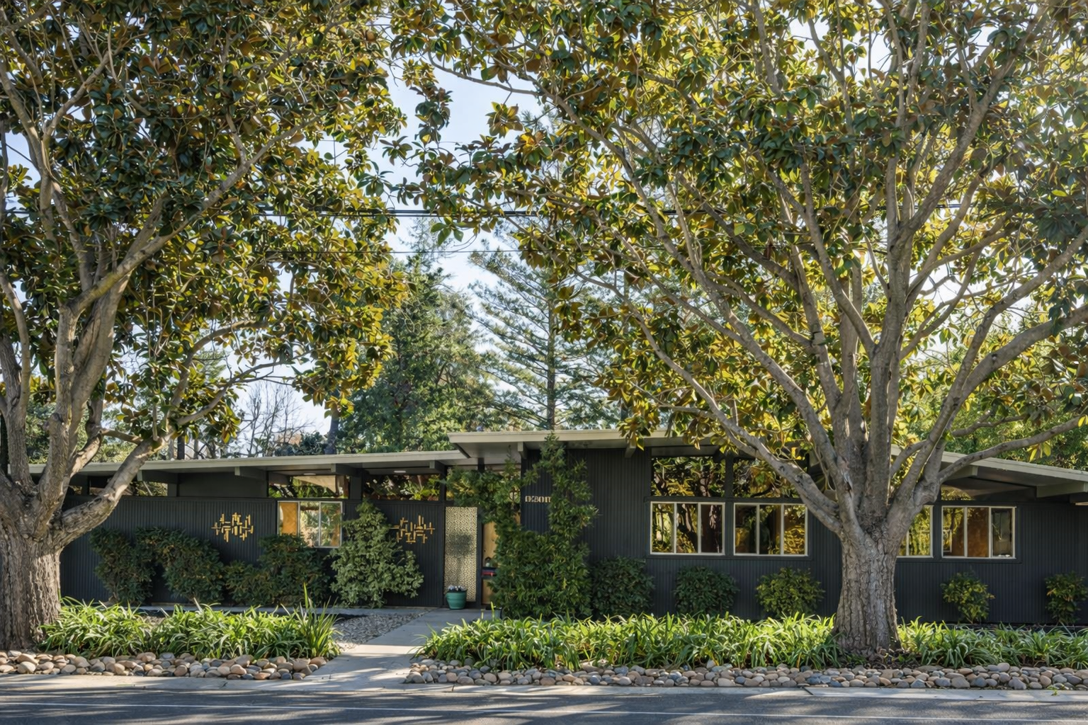 A mid-century modern house painted black with large windows, surrounded by trees and landscaped with rocks and green plants.