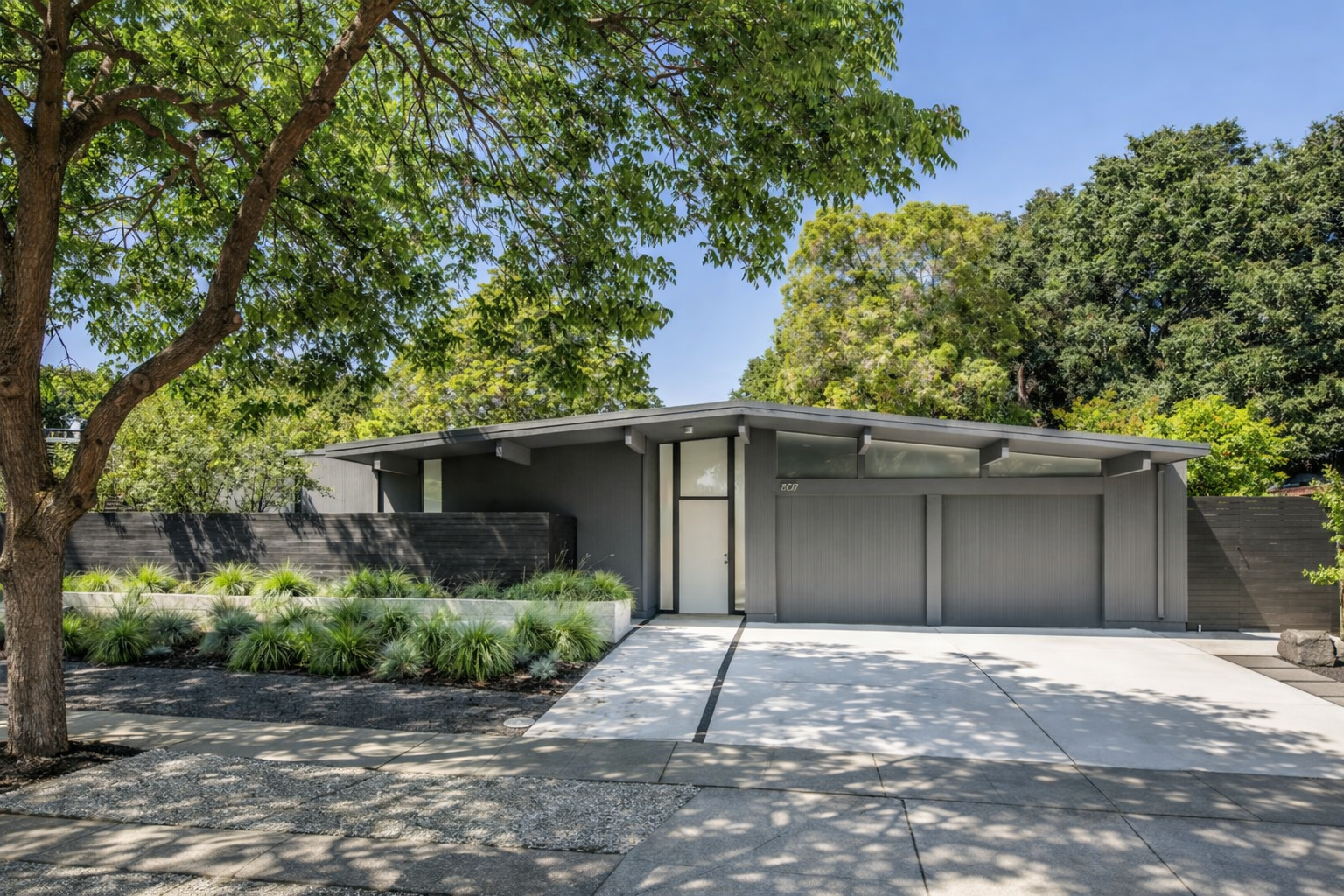 Modern gray house with sloped roof, surrounded by trees and greenery, with a concrete driveway in front.