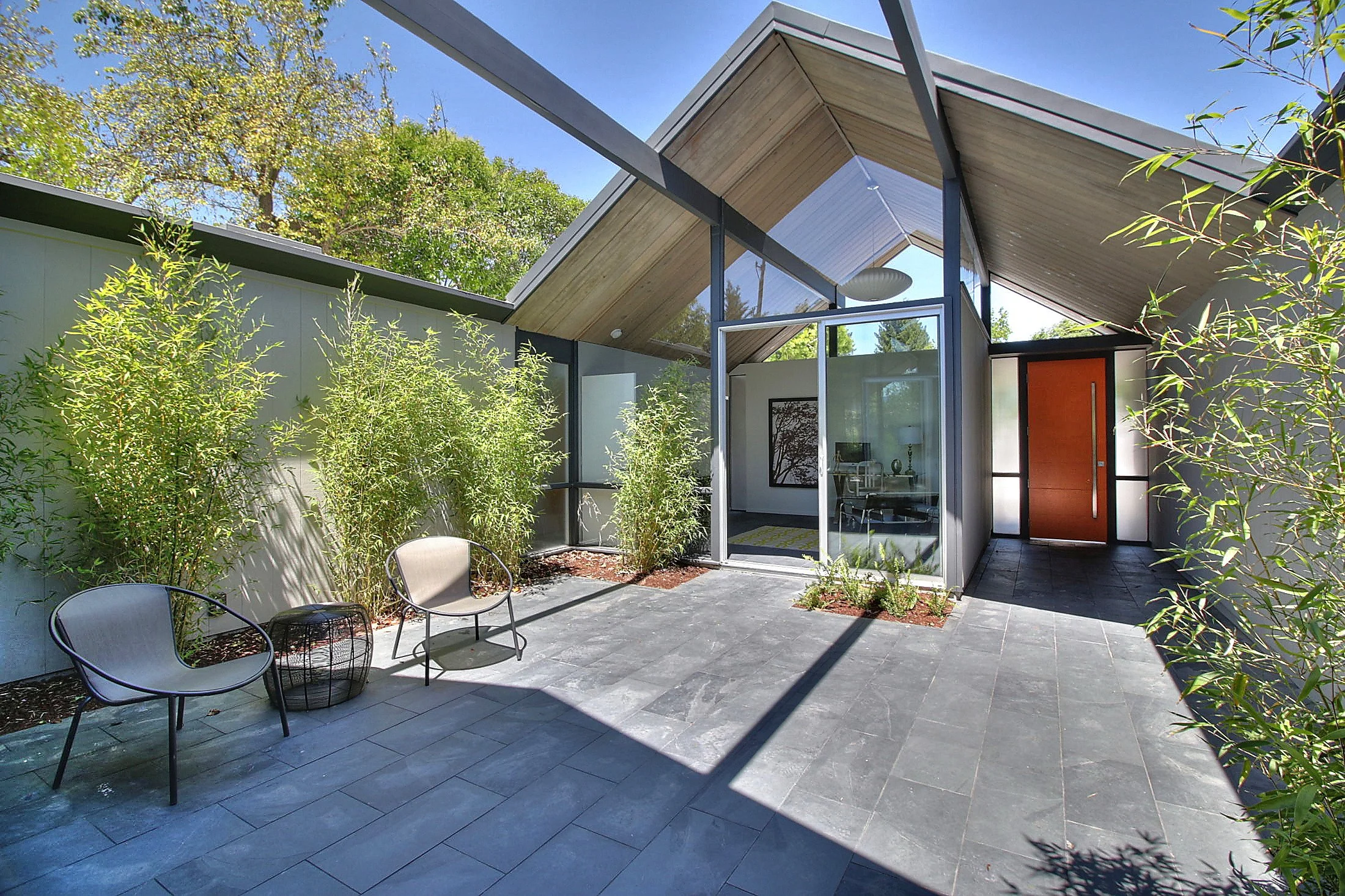 Modern house with large glass windows and a patio with two chairs and a small table, surrounded by green plants and trees under a sunny sky.