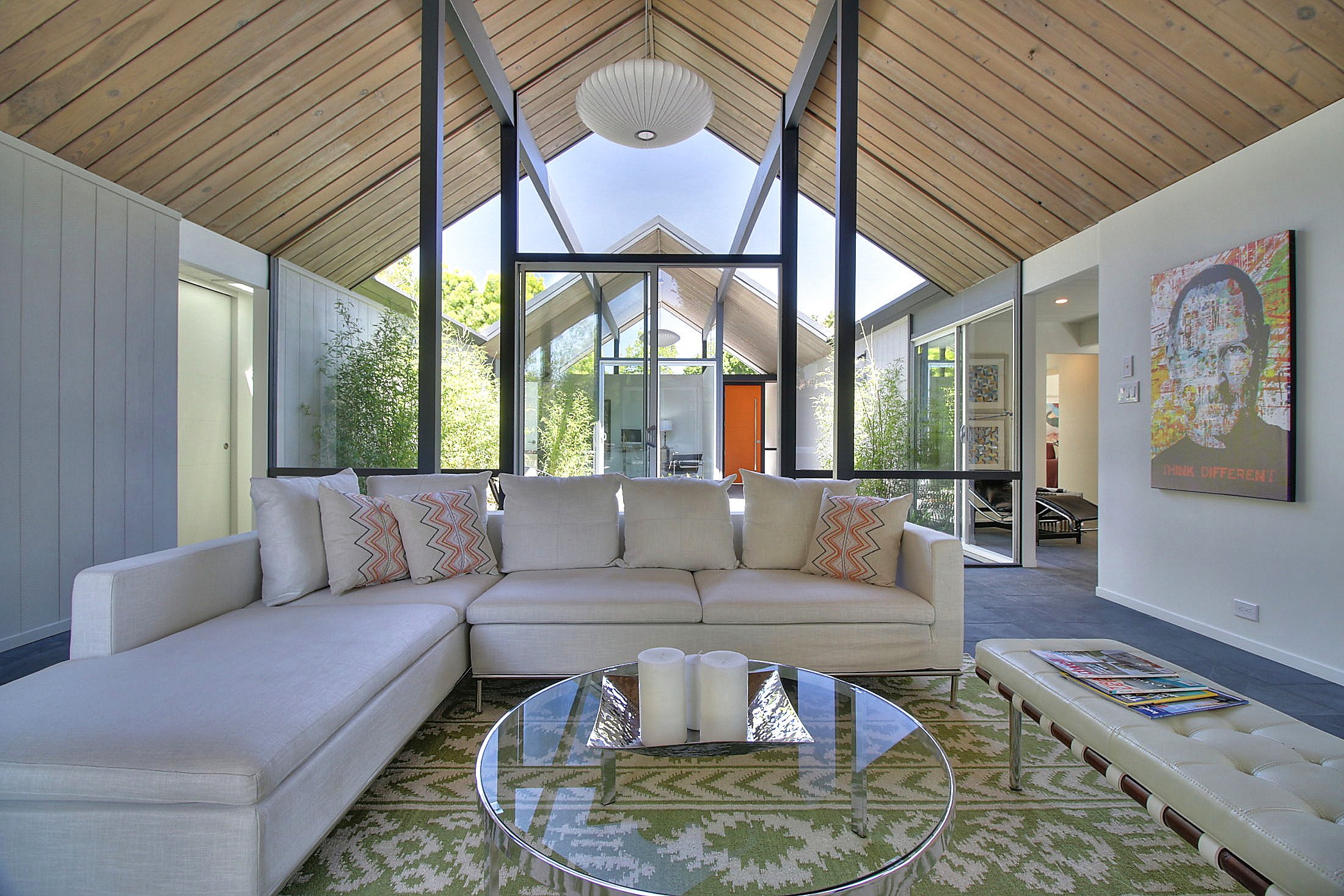 Modern living room with a white sectional sofa, colorful cushions, glass coffee table with candles, and large windows with a view of trees and outdoor space.
