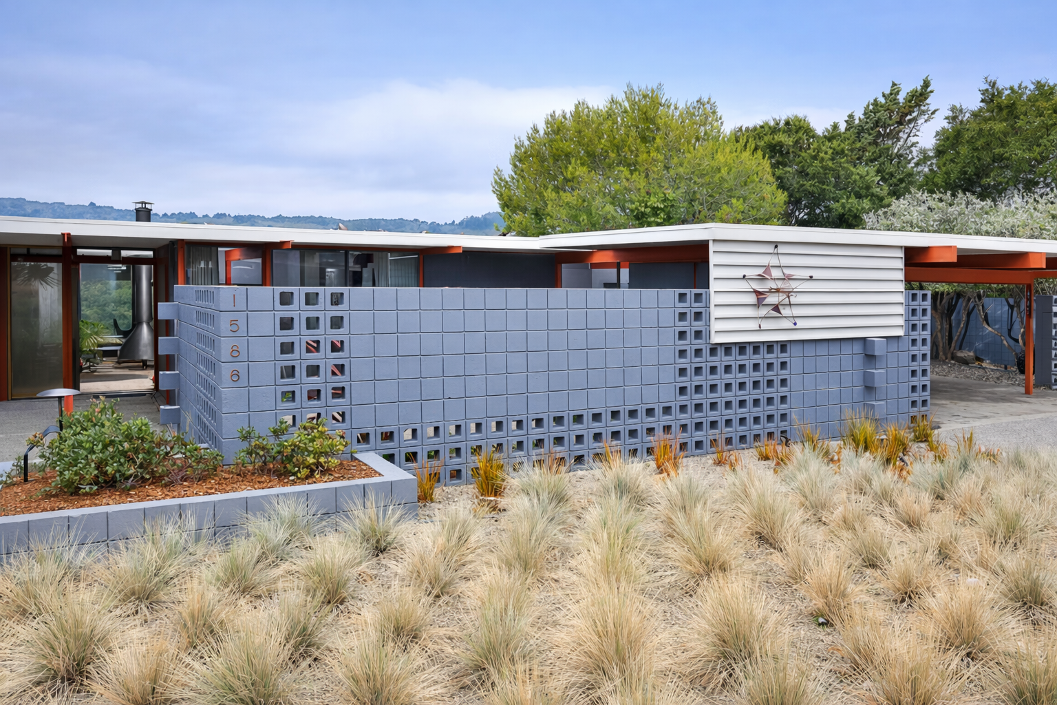 Modern house with a blue brick exterior, surrounded by desert vegetation and trees, under a cloudy sky.