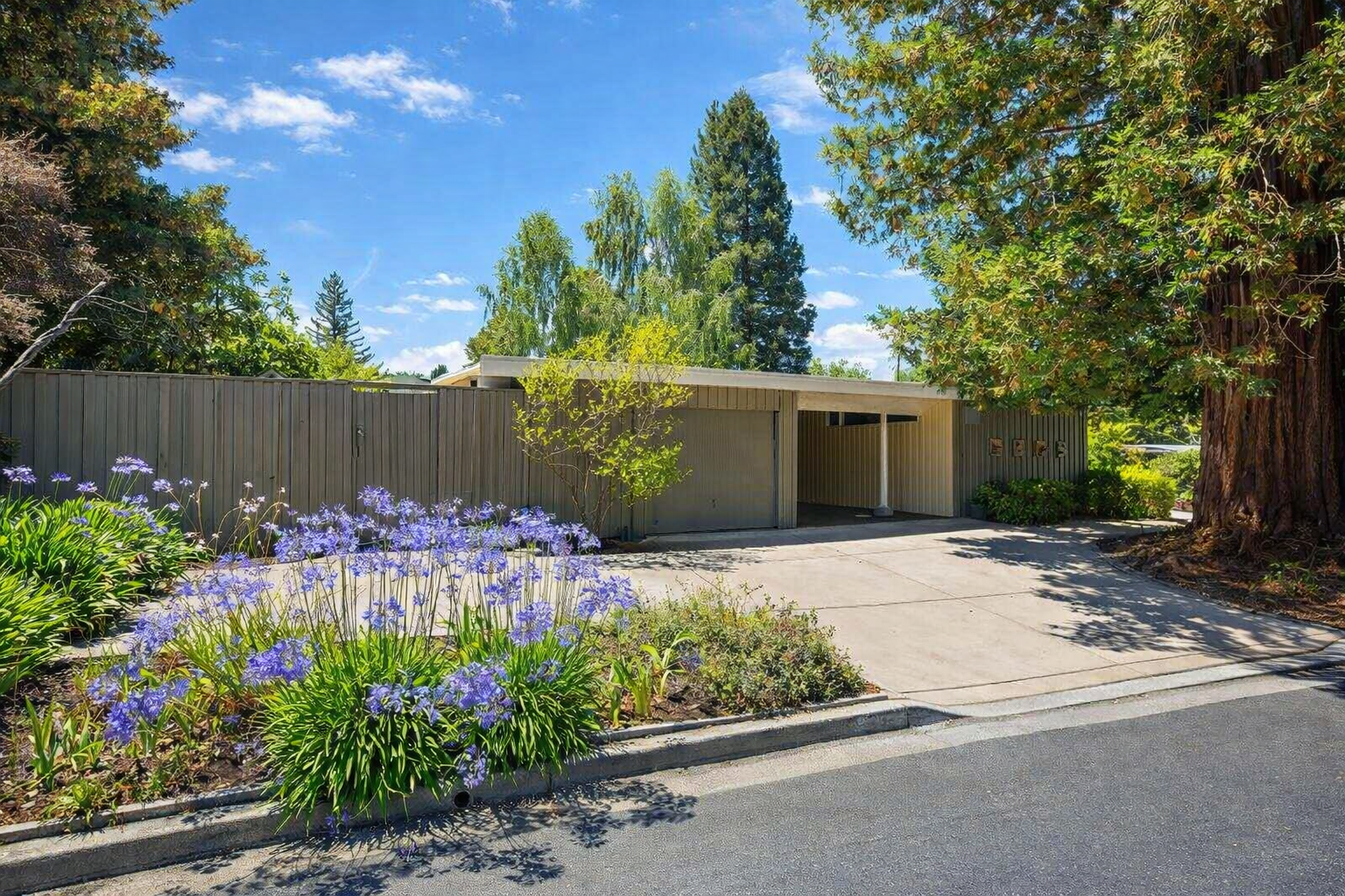 Residential house with a driveway, surrounded by green trees and purple flowers under a partly cloudy sky.