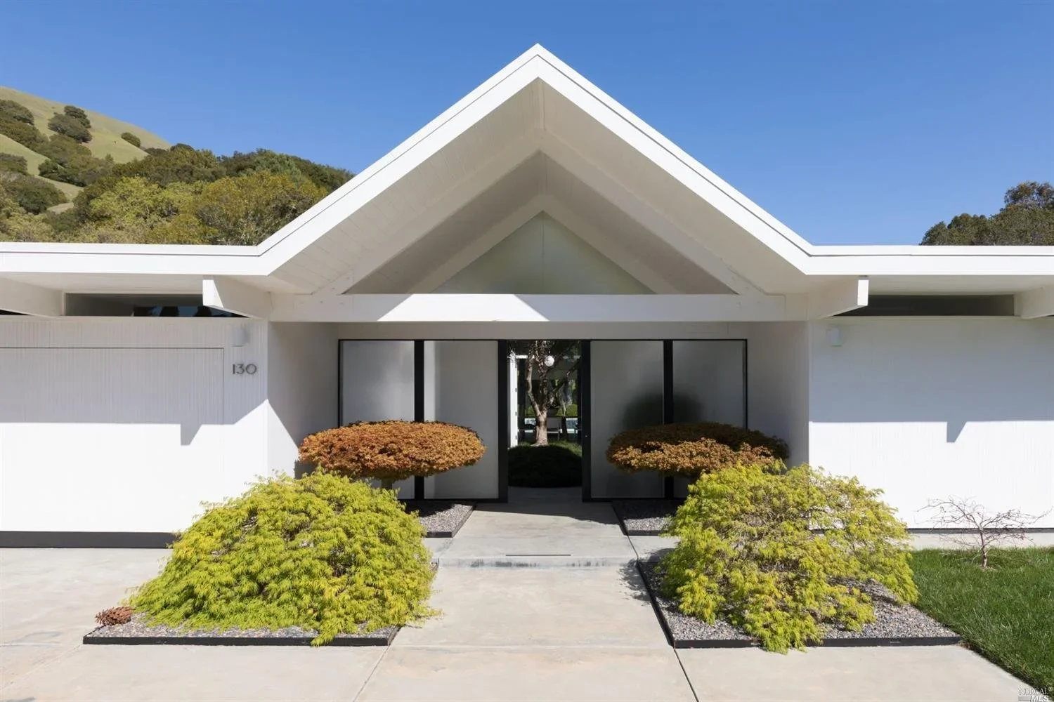 Front view of a modern white house with a peaked roof, glass doors, and landscaped bushes and trees in front.