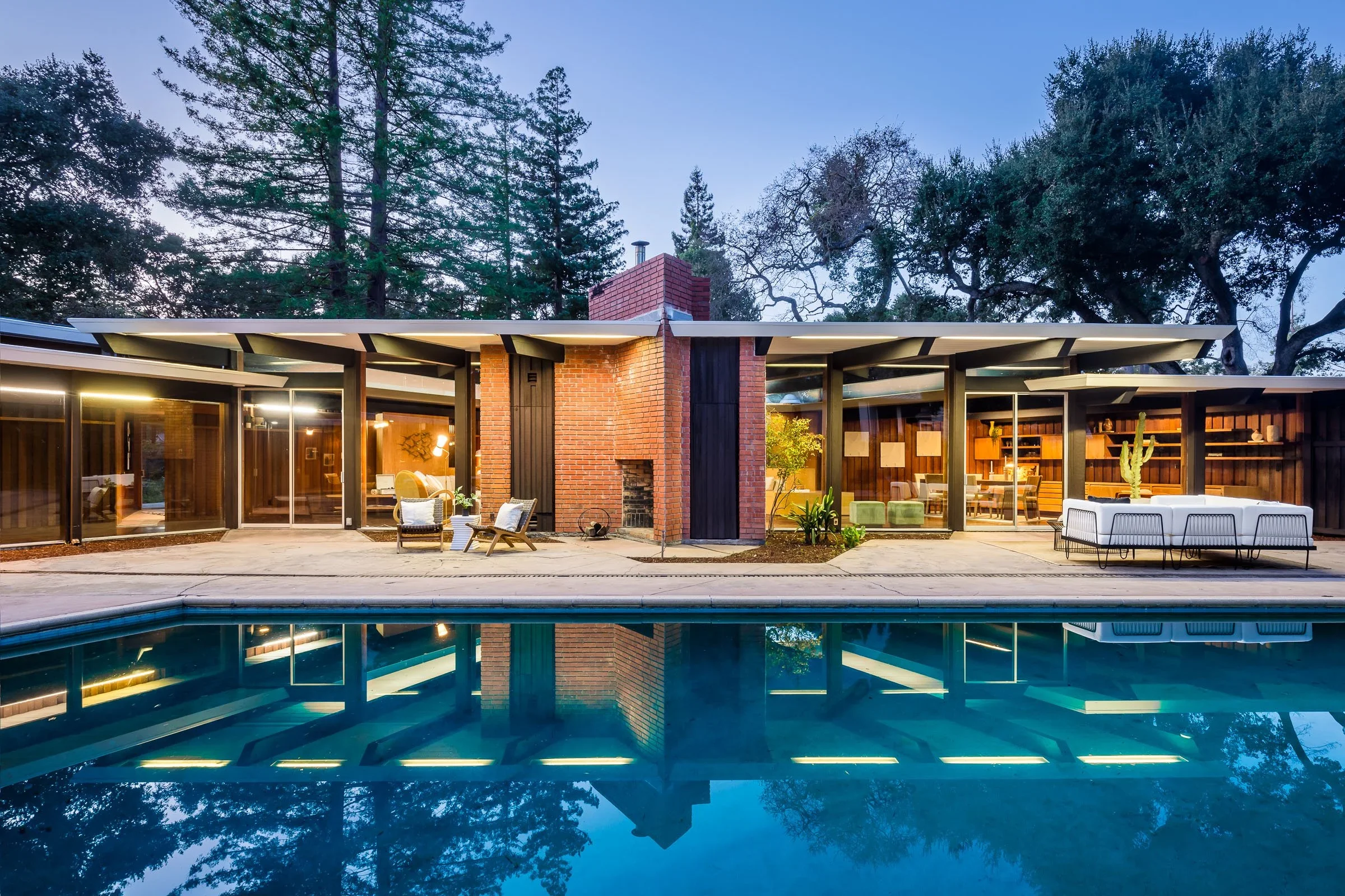 Modern house with large glass windows, brick chimney, and outdoor seating area, reflected in swimming pool, surrounded by trees at dusk.