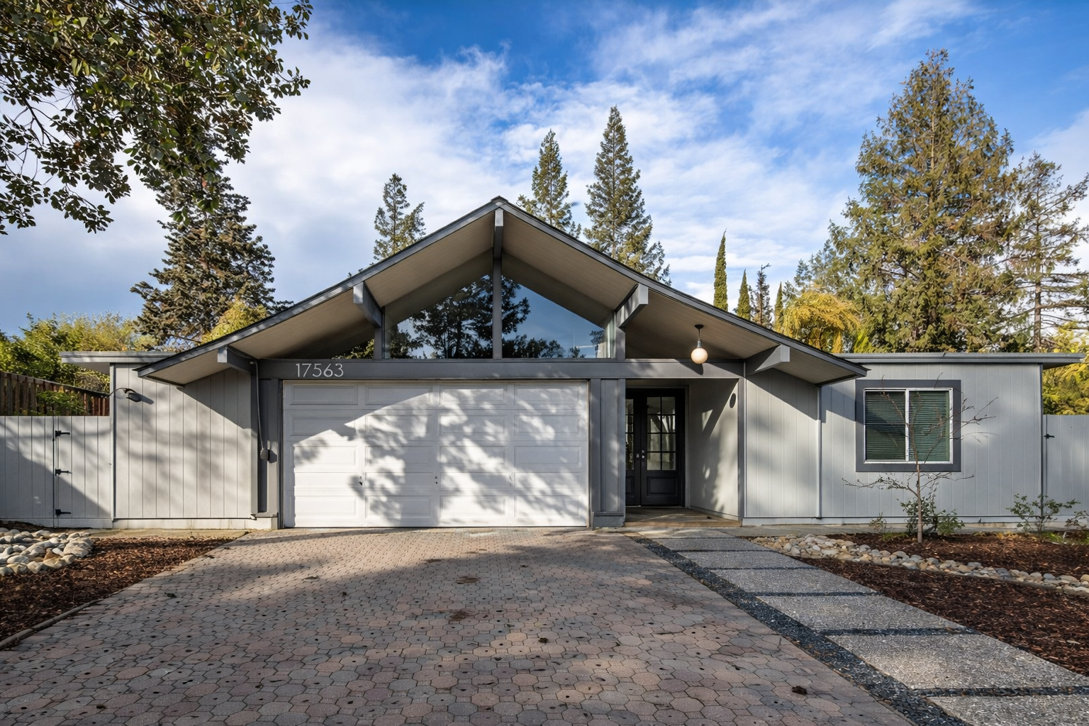Modern single-story house with a white exterior, dark trim, and a garage, surrounded by trees under a partly cloudy sky.