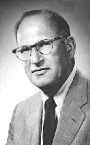 Black and white photo of a man wearing glasses, a suit, and a tie, looking serious.
