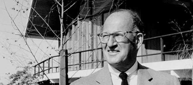 A black and white photo of a smiling man in glasses wearing a suit and tie, outdoors near a building with a railing and trees.
