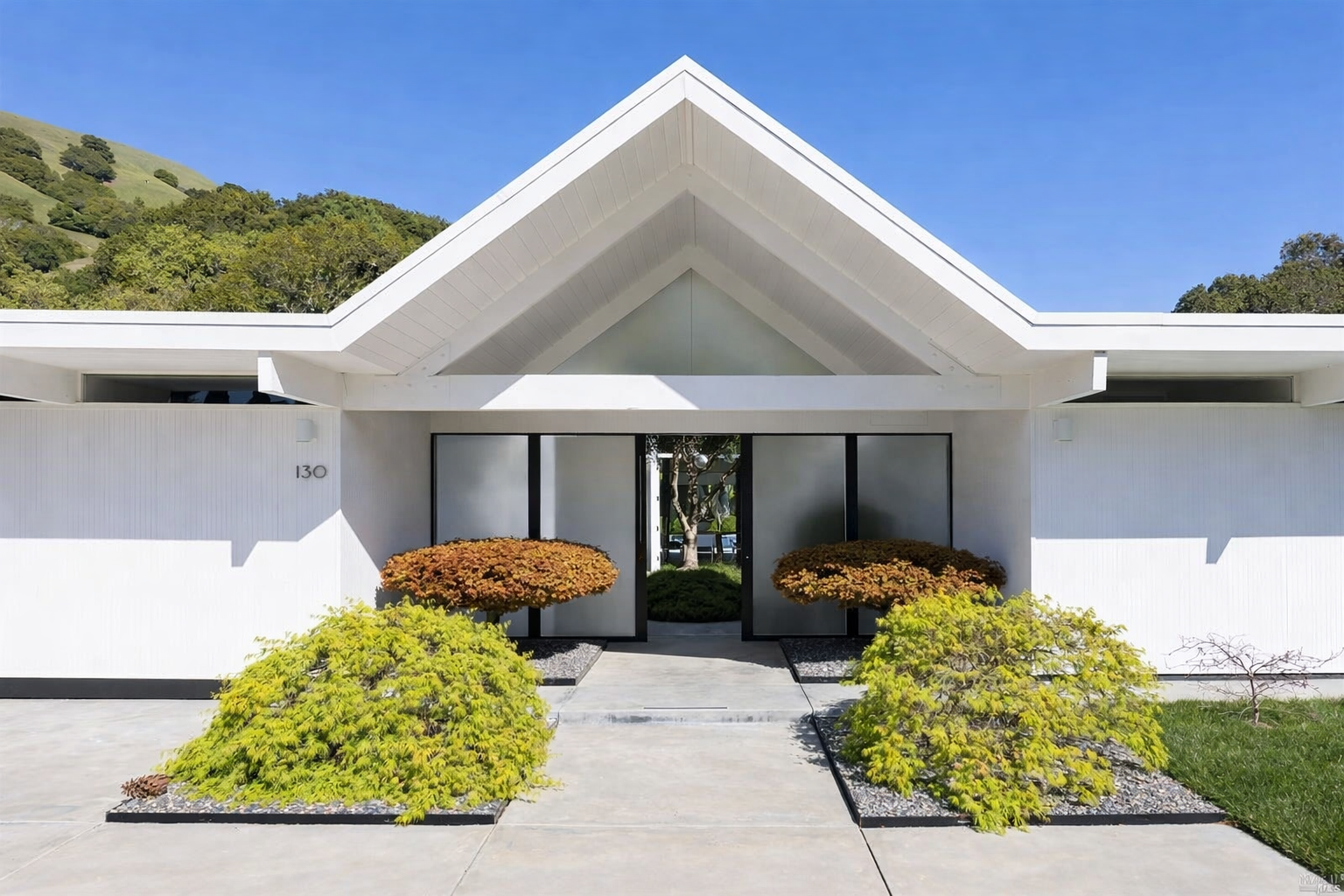 Front view of a modern white house with a peaked roof, glass front door, and landscaped bushes in front. Hills and trees are visible in the background under a clear blue sky.