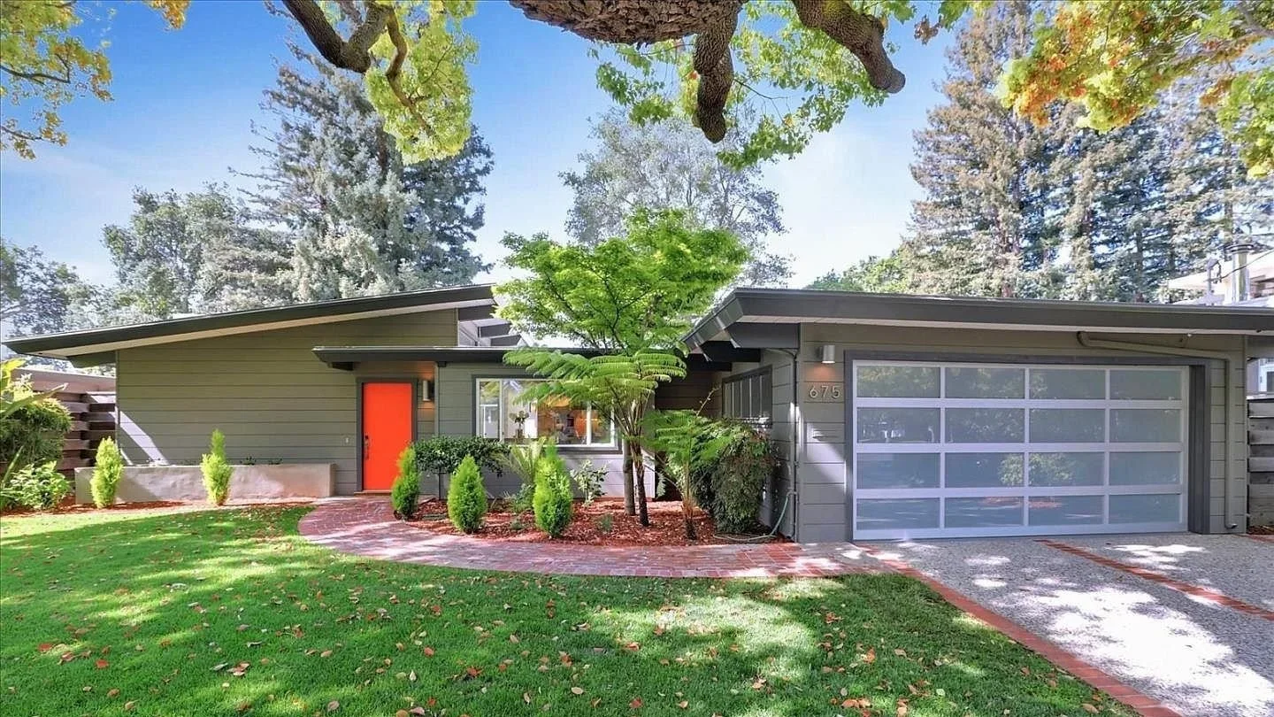 Front view of a modern house with a flat roof, gray siding, and a large garage door. The house features a bright orange front door, and a curved brick pathway leads to it. The yard is landscaped with grass, small bushes, and a tree in the front yard, with large trees in the background.