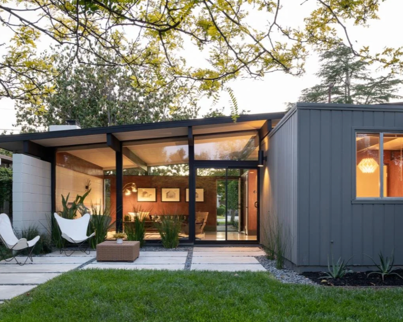 Modern house with large glass doors and windows, outdoor seating area with chairs and plants, green lawn, and trees in the background during evening.