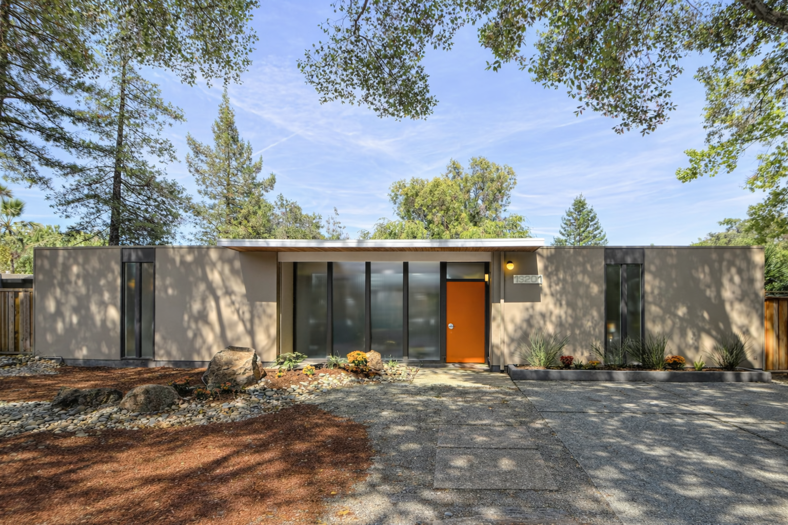 Modern single-story house with a flat roof, large glass windows, and a brown front door, surrounded by trees and natural landscaping.