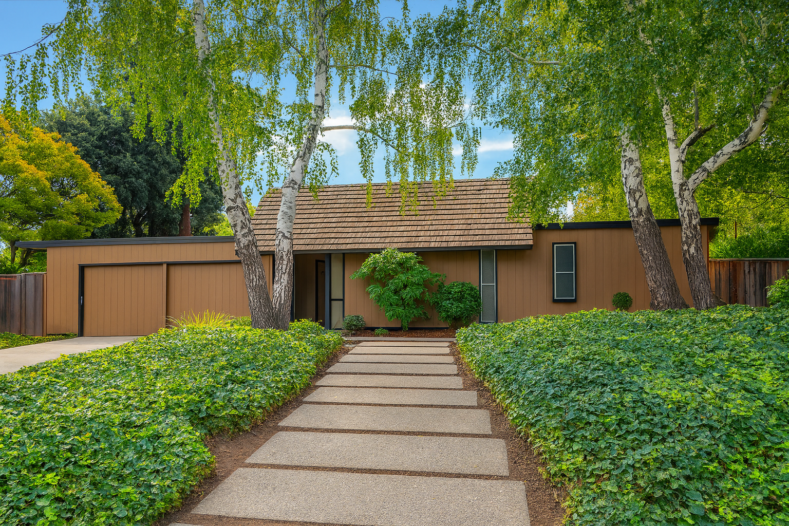 Modern house with a brown exterior, large windows, and a tiled roof, surrounded by green trees and bushes, with a concrete pathway leading to the entrance.